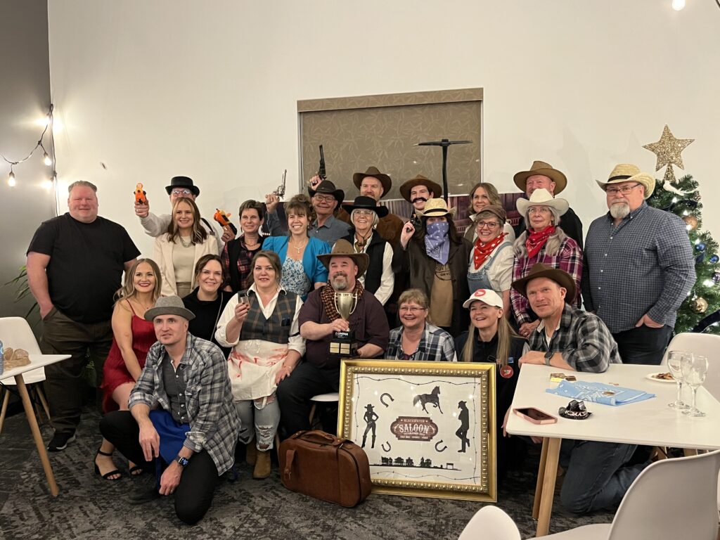 A company holiday party gathers together for a group photo. Everyone is smiling and donning wild west apparel.